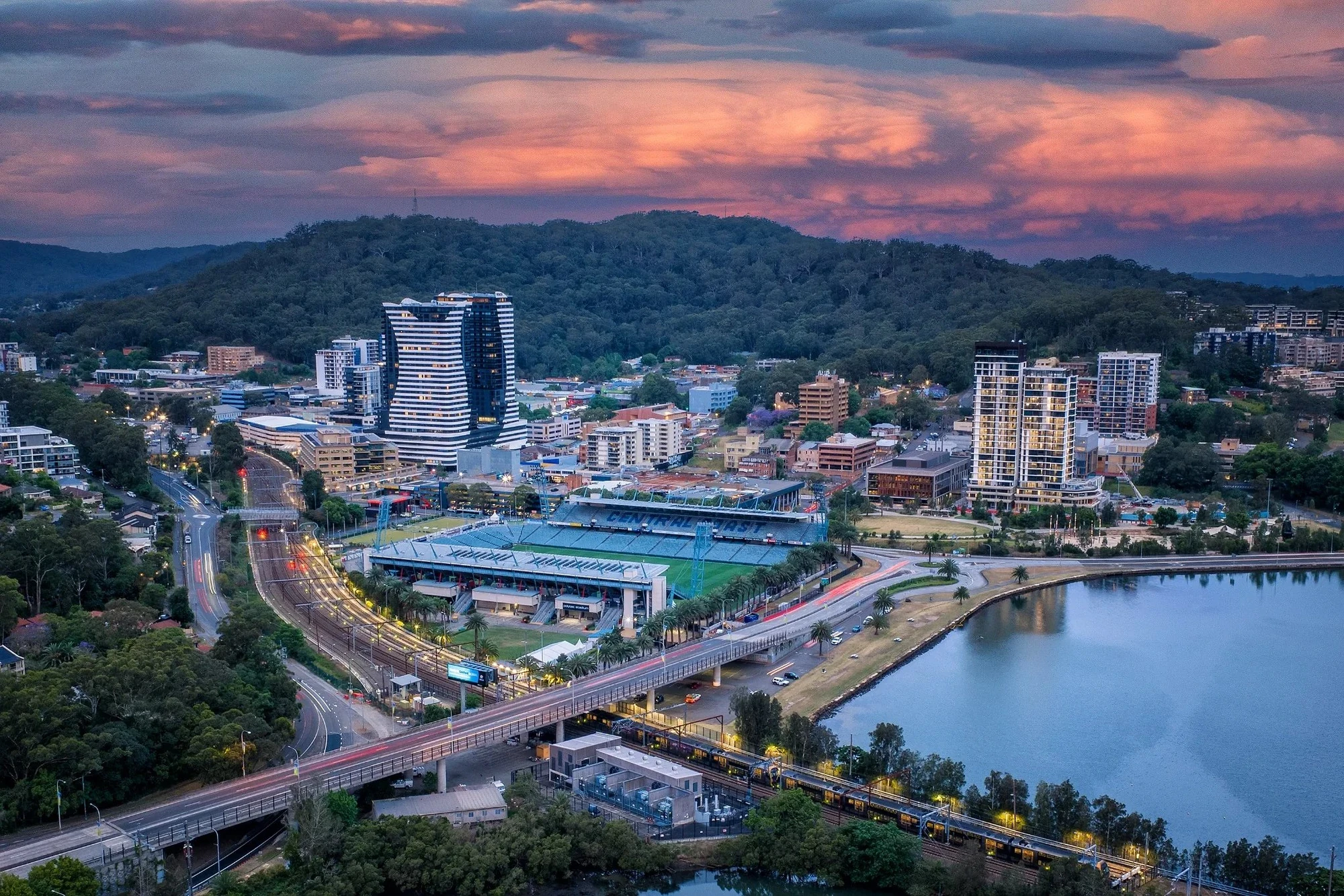Aerial sunset view of Gosford CBD showing Central Coast Stadium, high-rise buildings, and Brisbane Water surrounded by bushland