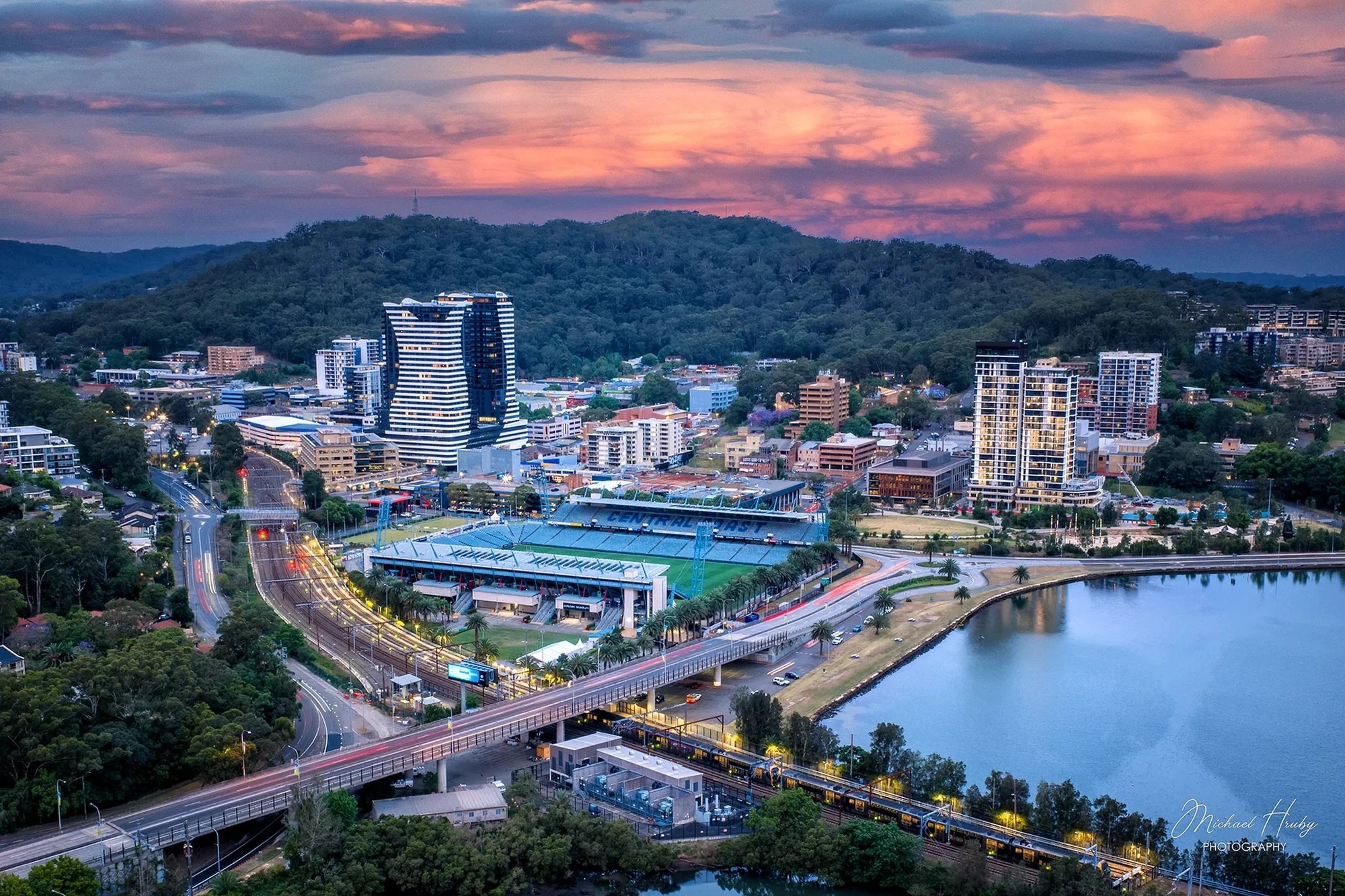 Aerial sunset view of Gosford CBD showing Central Coast Stadium, high-rise buildings, and Brisbane Water surrounded by bushland