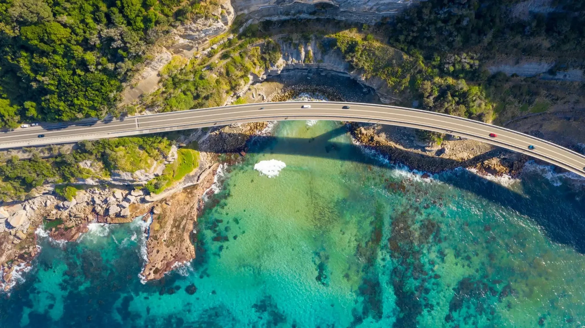 Aerial view of the Sea Cliff Bridge curving over turquoise water on the NSW coast, surrounded by cliffs and natural greenery