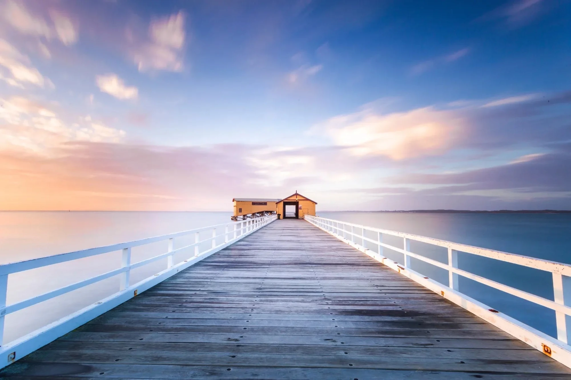 Long wooden jetty stretching into calm water at sunrise, with soft clouds and warm pastel light above the horizon