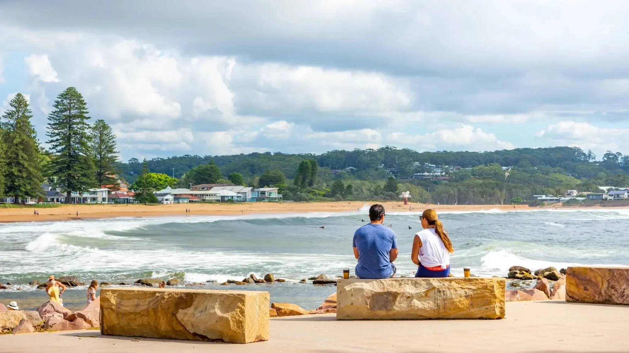 Two people sitting along the Avoca Beach promenade looking out over the waves on a cloudy day, Central Coast NSW