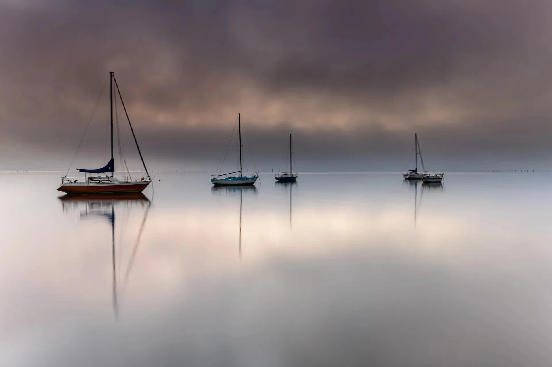 Still water reflecting muted skies as four sailboats rest at anchor on the Central Coast