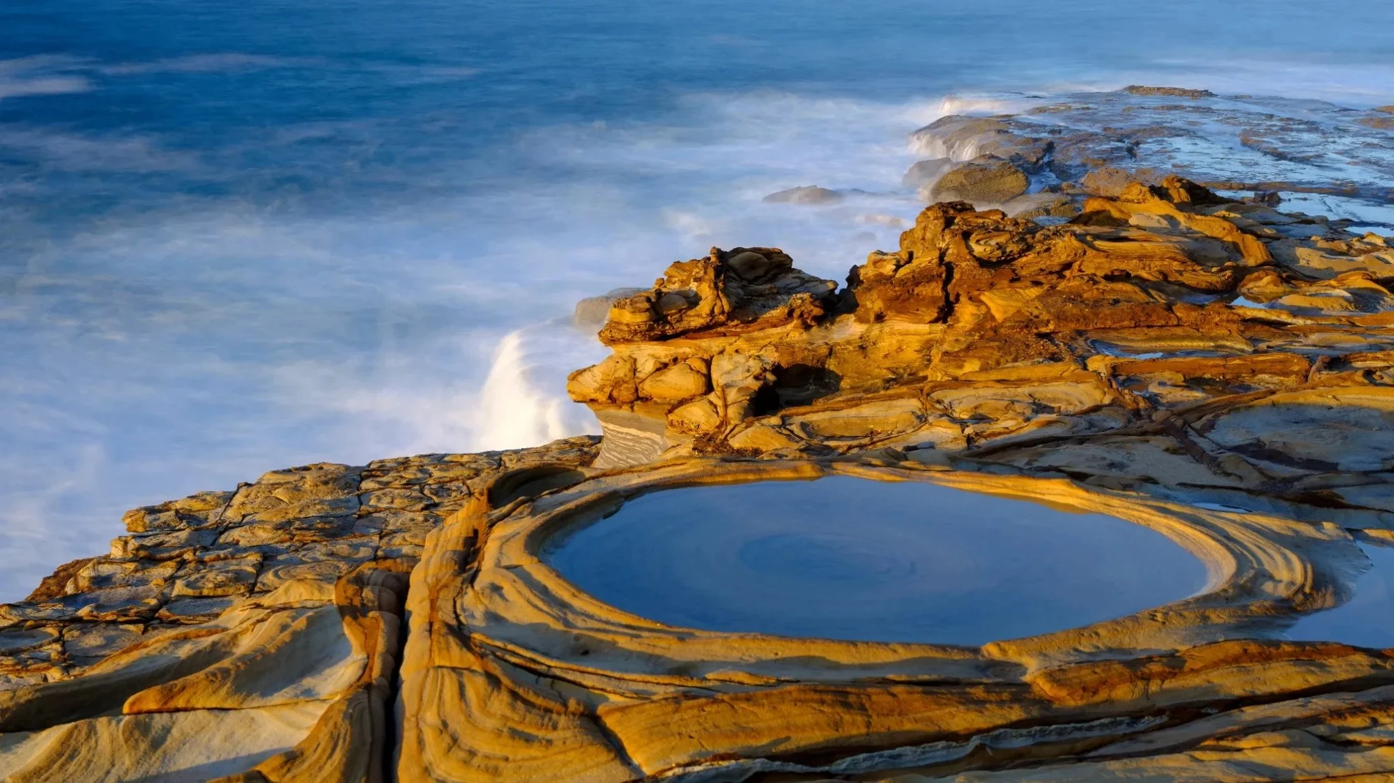 Sunrise lighting the layered sandstone rock pools along the Bouddi National Park coastline on the Central Coast, with soft waves creating a calm, grounding coastal scene